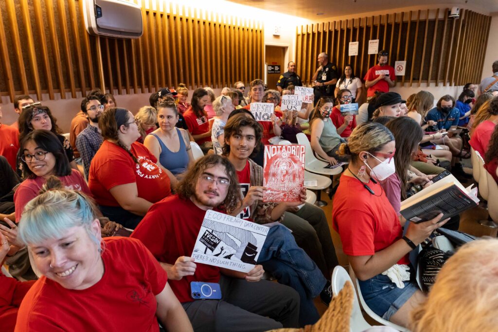 A group of people in red hold signs while sitting in a committee meeting